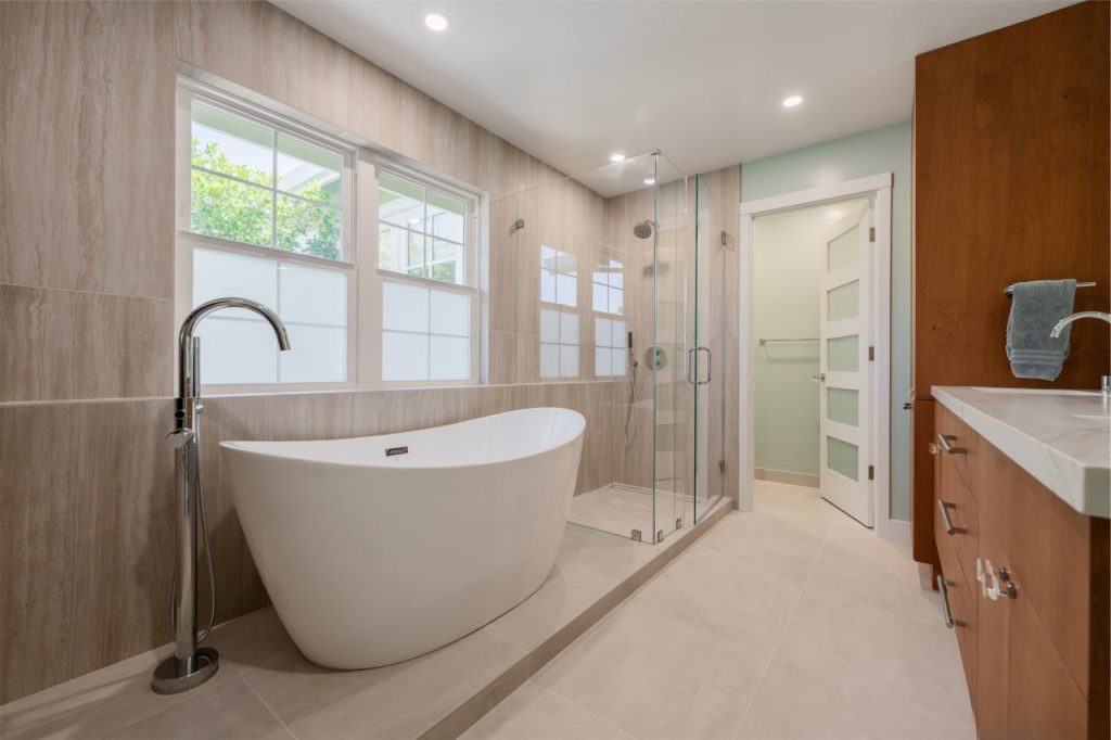 Elegant freestanding tub positioned near frameless glass shower with white contemporary vanity and natural light.