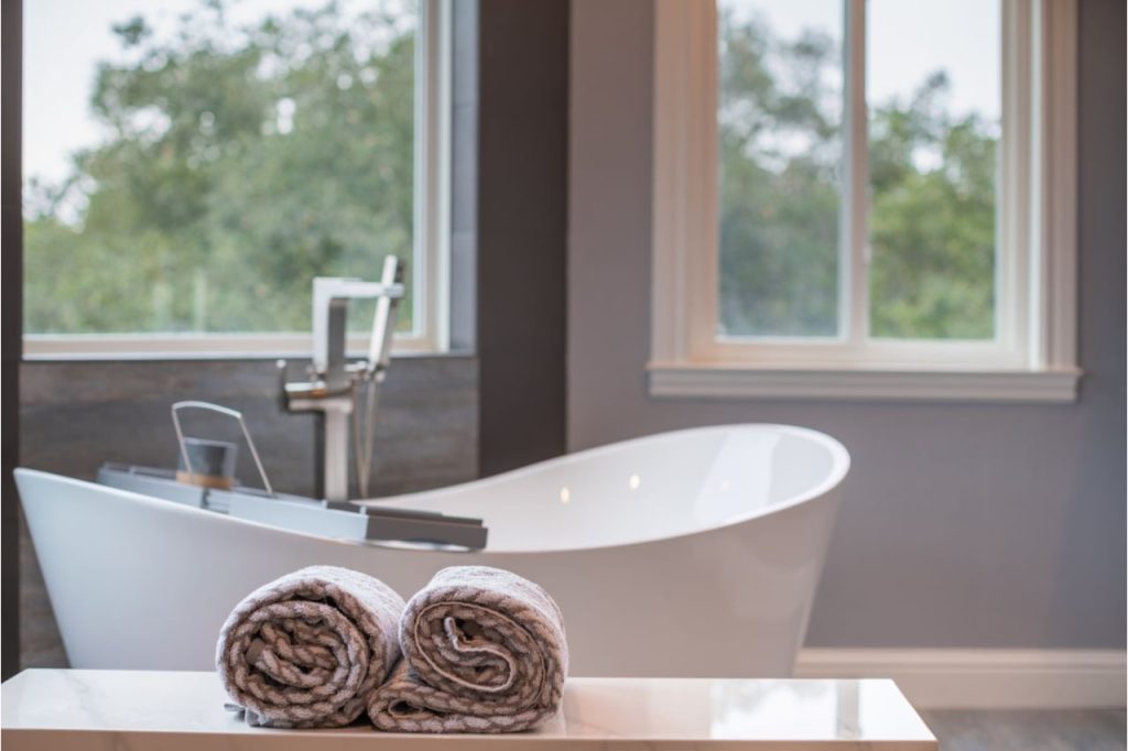 Elegant freestanding tub positioned beside large window with rolled towels and black contemporary vanity cabinetry.