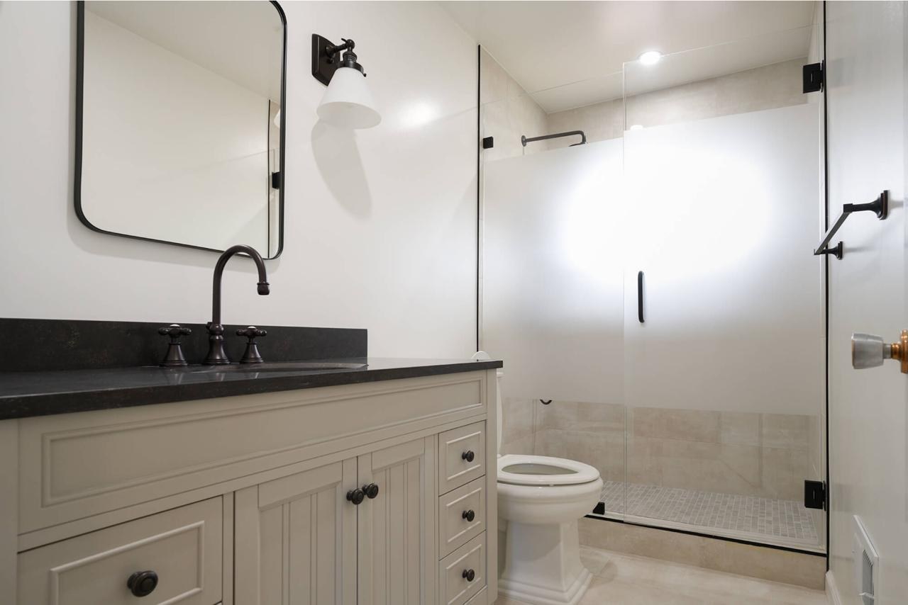 Bathroom after: frameless glass shower with matte black hardware with dark stone countertop and painted inset cabinetry, by ASL Remodeling.