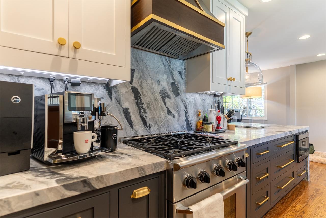 Modern kitchen renovation featuring white upper cabinets with gold hardware, dark lower cabinets, dramatic grey and white marble backsplash, stainless steel range hood, gas cooktop, and wood-look countertop