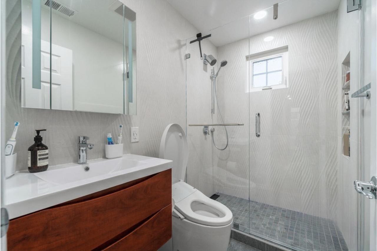 Luxury bathroom remodel featuring large-format gray porcelain tile, wood-tone floating vanity with undermount sinks, frameless glass shower enclosure, and modern pendant lighting fixtures