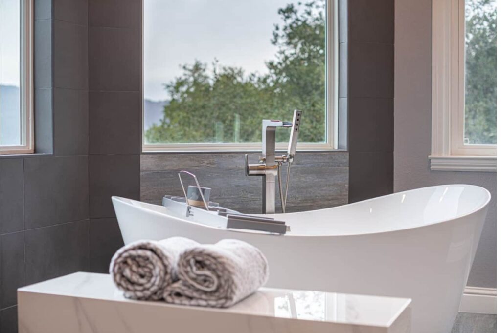 Freestanding soaking tub with floor-mounted filler and handheld sprayer set beneath windows overlooking trees.
