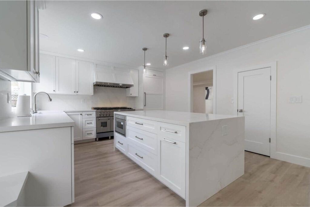 All-white kitchen with a waterfall-edge island, pendant lighting, and a stainless steel range beneath a vent hood.
