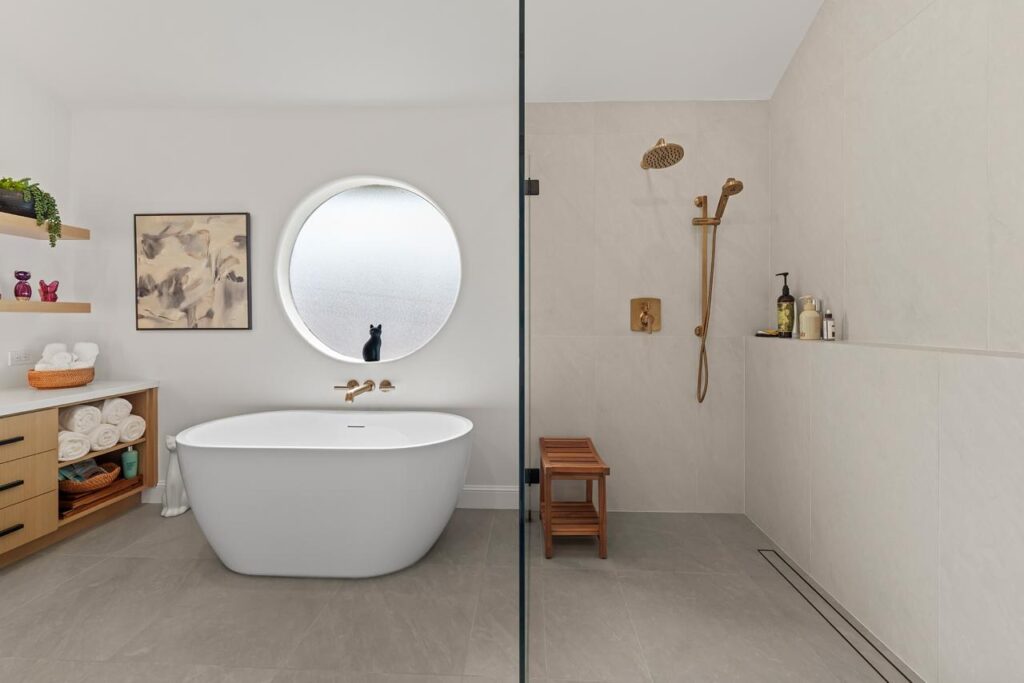 Primary bathroom after: freestanding soaking tub with round window with white soaking tub and brushed brass fixtures, by ASL Remodeling.